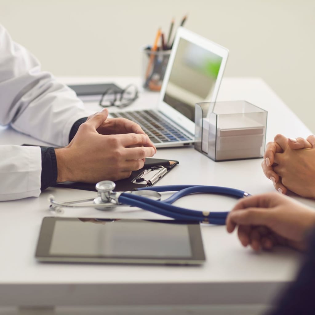 During a primary care consultation, the doctor and patient are seated at a desk, surrounded by a laptop, stethoscope, tablet, and a box of tissues.